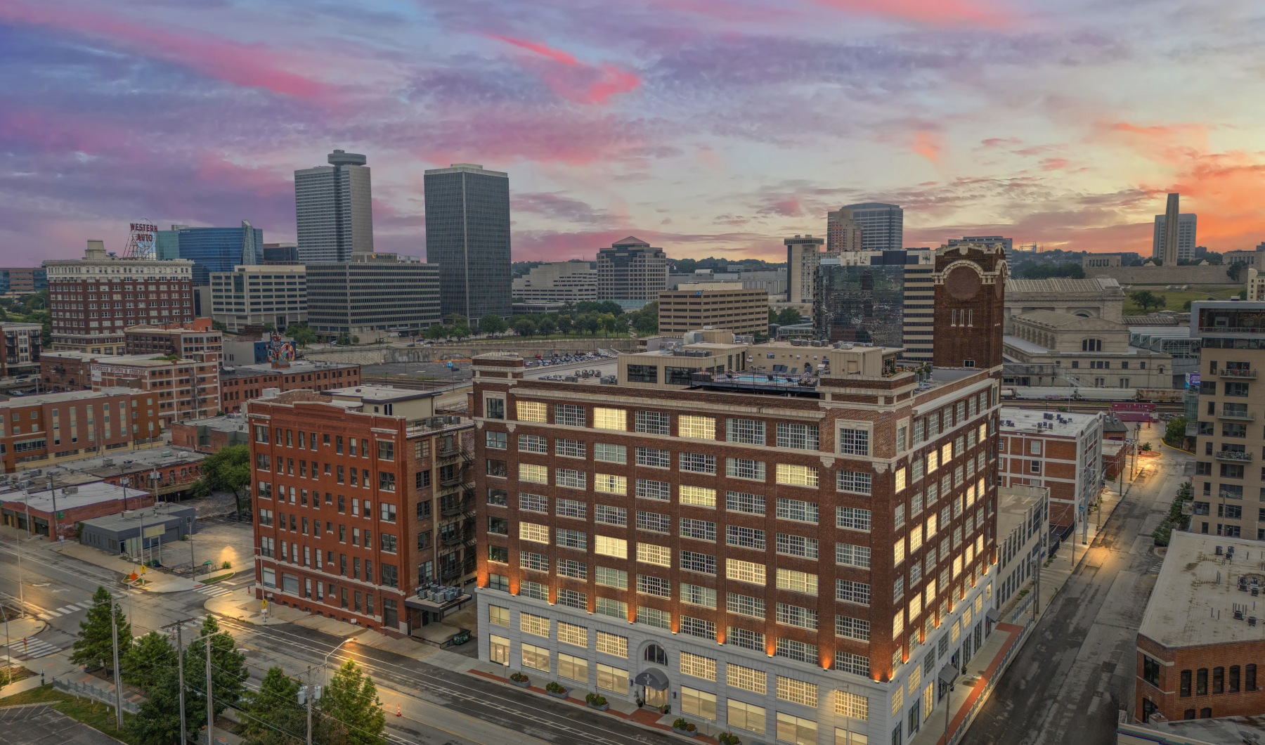 Exterior view of Piper Lofts in Kansas City featuring illuminated brick architecture against a vibrant sunset city skyline