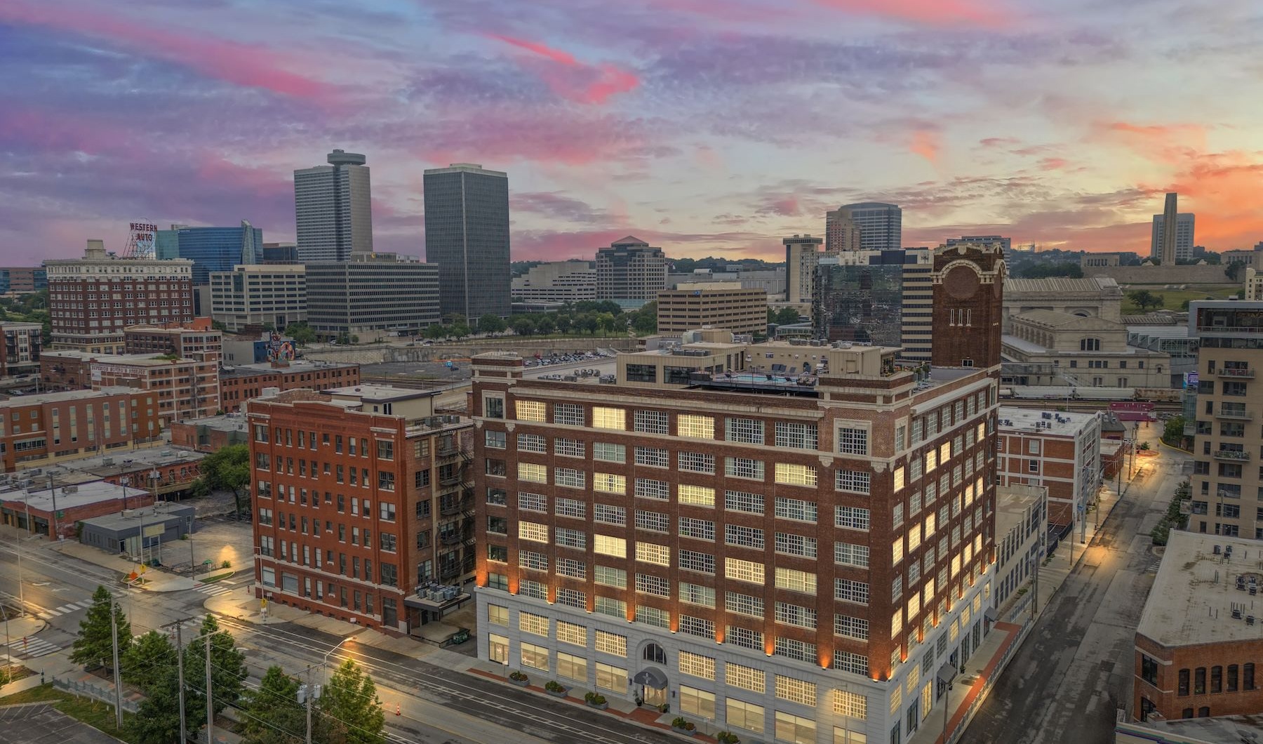 Exterior view of Piper Lofts in Kansas City featuring illuminated brick architecture against a vibrant sunset city skyline